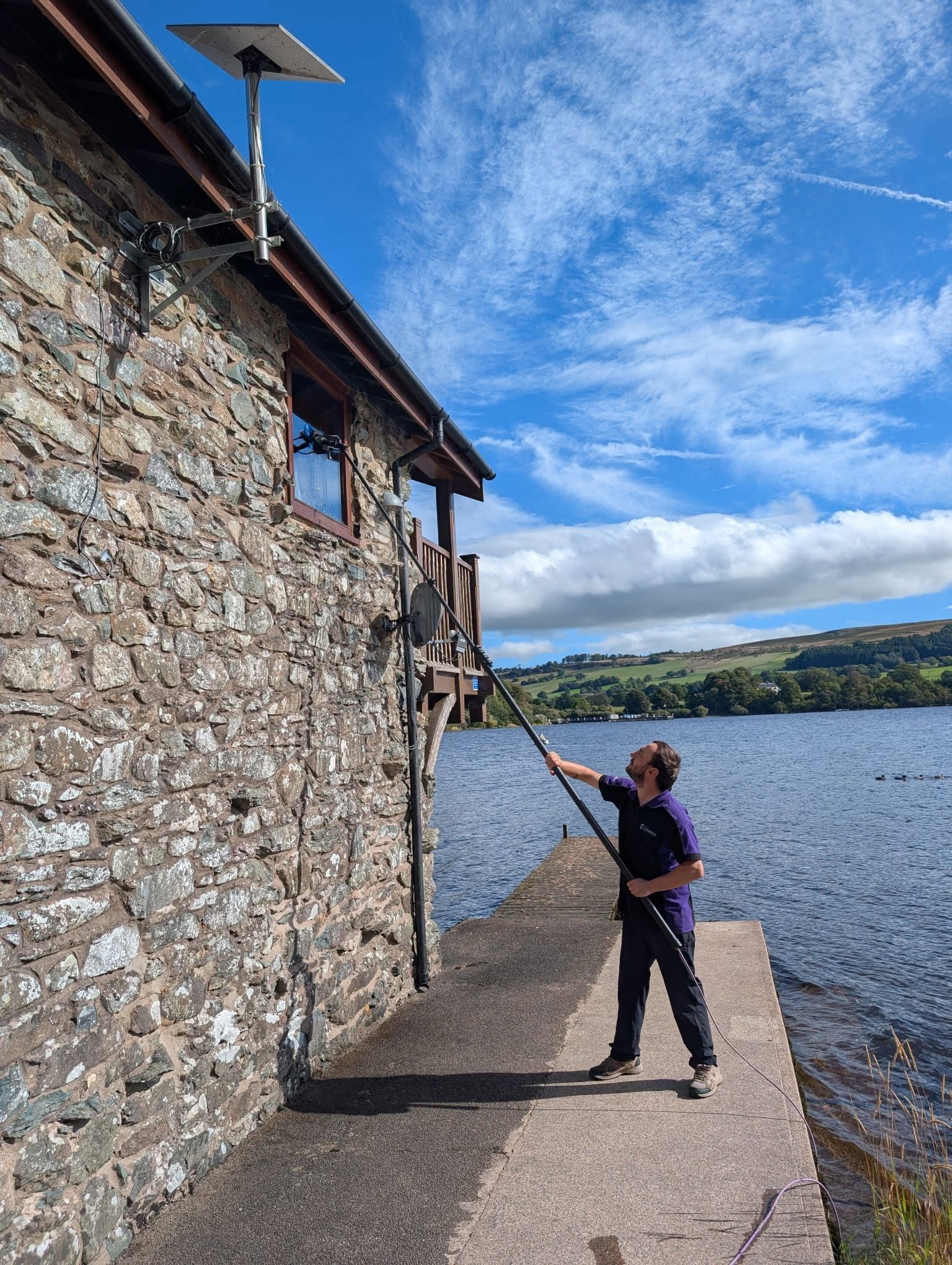 Cleaning the windows of a stone building from a lakeside jetty, with fells behind