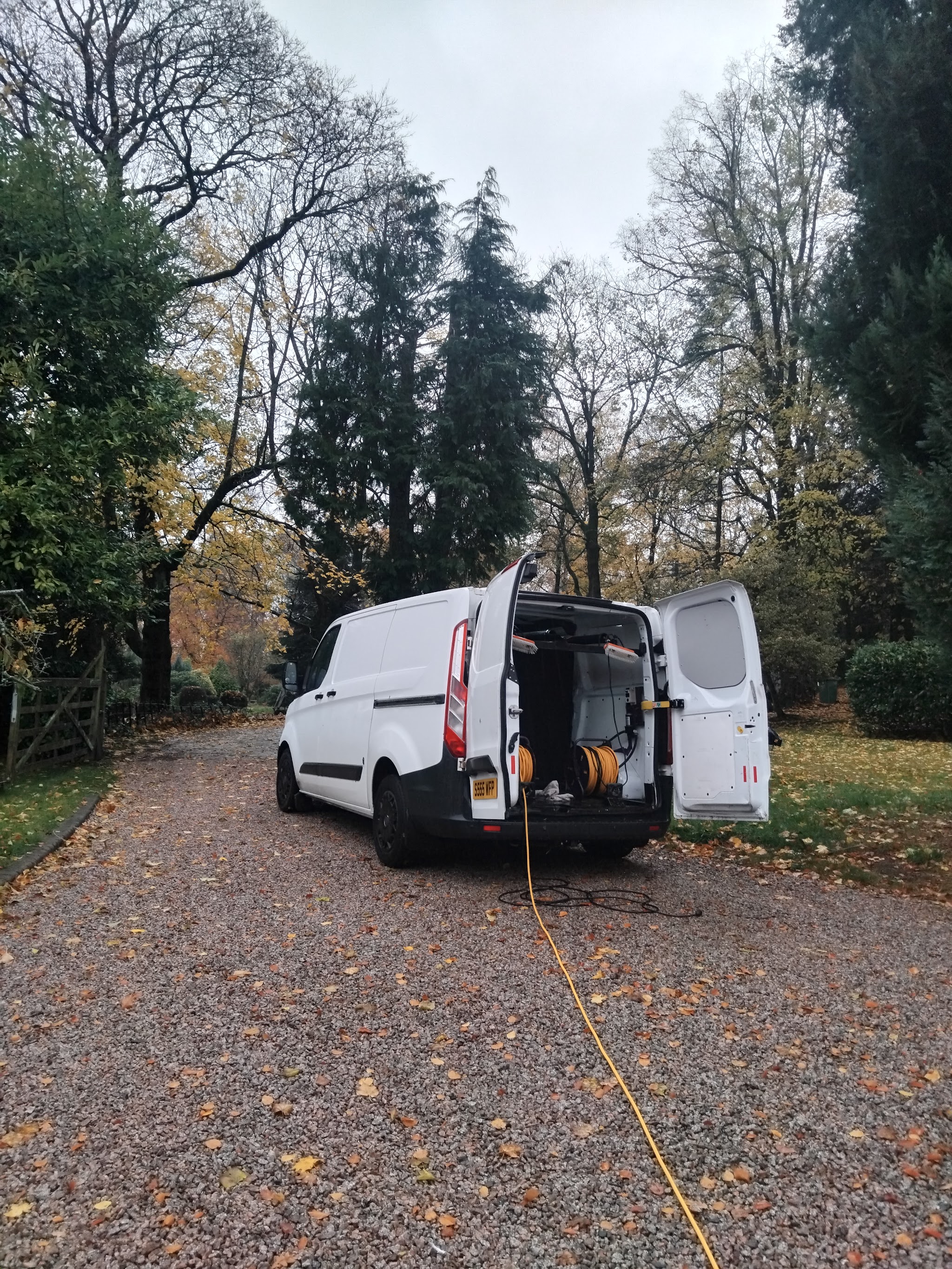 The van with its rear doors open showing the hose reel, on a gravel drive among autumn trees